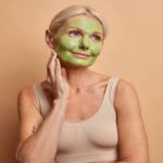 Portrait of dreamy middle aged woman applies green mask on face stands thoughtfully and looks away undergoes beauty procedures dressed casually isolated over beige background. Skin care concept
